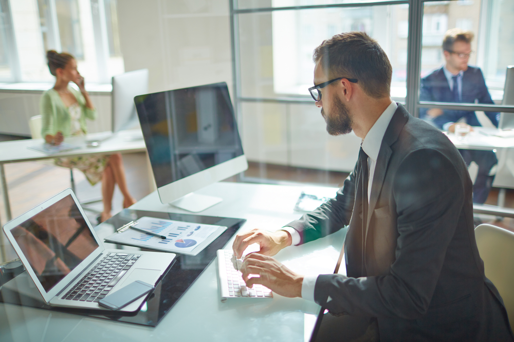 homem de terno e óculos mexendo no computador e com mesa de trabalho à vista