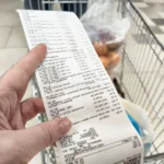 Mão segurando cupom fiscal longo em frente a um carrinho de supermercado, ilustrando a aplicação do NCM 2025 em notas fiscais.